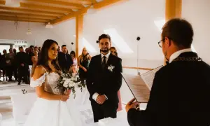 Para młoda podczas ceremonii cywilnej. Panna młoda w białej sukni, pan młody w garniturze. Goście obserwują. Czy po ślubie cywilnym można być chrzestnym?