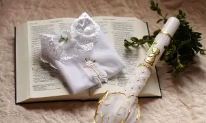 Biała szata, świeca z aniołkiem i gałązka symbol chrztu na otwartej Biblii.