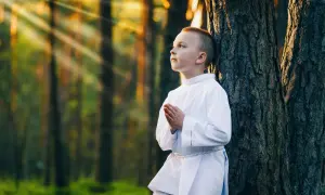 Chłopiec w albie modli się w lesie, promienie słońca przebijają się przez drzewa. Piękne sesje komunijne w plenerze.