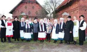 Grupa osób w tradycyjnych strojach wielkopolskich pozuje przed ceglanym budynkiem.