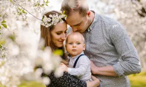 Urocza sesja urodzinowa w plenerze. Rodzice czule obejmują swoje dziecko wśród kwitnących drzew.
