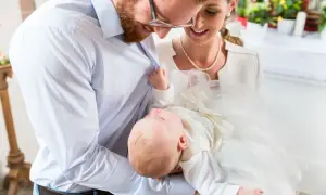 Szczęśliwi rodzice trzymają niemowlę w białej szatce. W tle kościół, gdzie odbywają się chrzciny dziecka. Przesądy związane z tym wydarzeniem są różne.