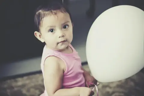 Urocza dziewczynka w różowej bluzeczce podczas sesji na roczek w domu, trzymająca biały balon.
