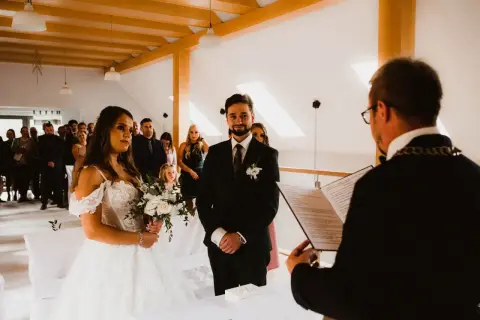 Para młoda podczas ceremonii cywilnej. Panna młoda w białej sukni, pan młody w garniturze. Goście obserwują. Czy po ślubie cywilnym można być chrzestnym?
