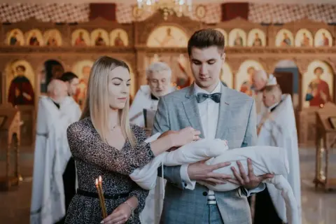 Młodzi rodzice trzymają dziecko do chrztu prawosławnego. W tle widać cerkiew i duchownych.