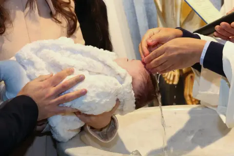 Chrzest święty: ksiądz polewa głowę dziecka wodą. Rodzice trzymają maluszka w białym kocyku.