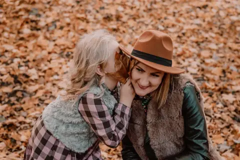 Córka szepcze mamie sekret do ucha podczas sesji mama córka wśród jesiennych liści.