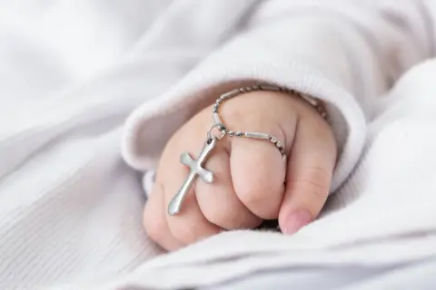 Dziecięca dłoń z krzyżykiem na bransoletce, symbol błogosławieństwa. Planujesz jak zorganizować chrzciny?