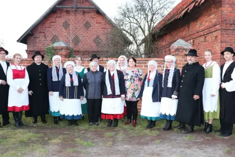 Grupa osób w tradycyjnych strojach wielkopolskich pozuje przed ceglanym budynkiem.