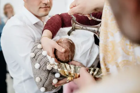 Chrzest dziecka podczas chrzcin. Woda z ozdobnego dzbanka spływa na główkę niemowlęcia.