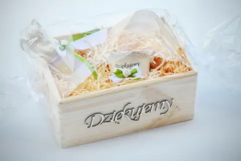 Drewniana skrzynka z napisem "Dziękujemy", wypełniona siankiem, zawiera filiżankę z napisem "Dziękujemy" i ozdobne wstążki. Idealny tekst podziękowania dla księdza.