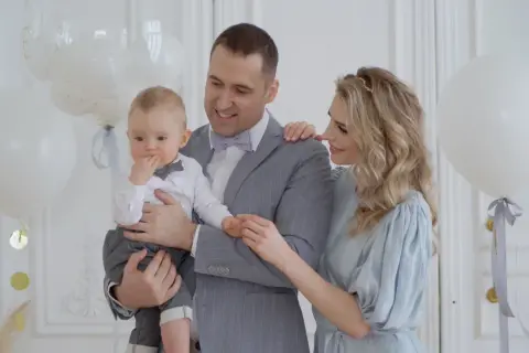 Uśmiechnięci rodzice trzymają synka w eleganckim stroju. Białe balony i złote konfetti tworzą atmosferę idealną na chrzciny dla chłopca.
