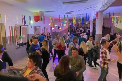 Wesoła zabawa w remizie, ludzie tańczą przy muzyce. Kolorowe serpentyny i balony tworzą świąteczną atmosferę.