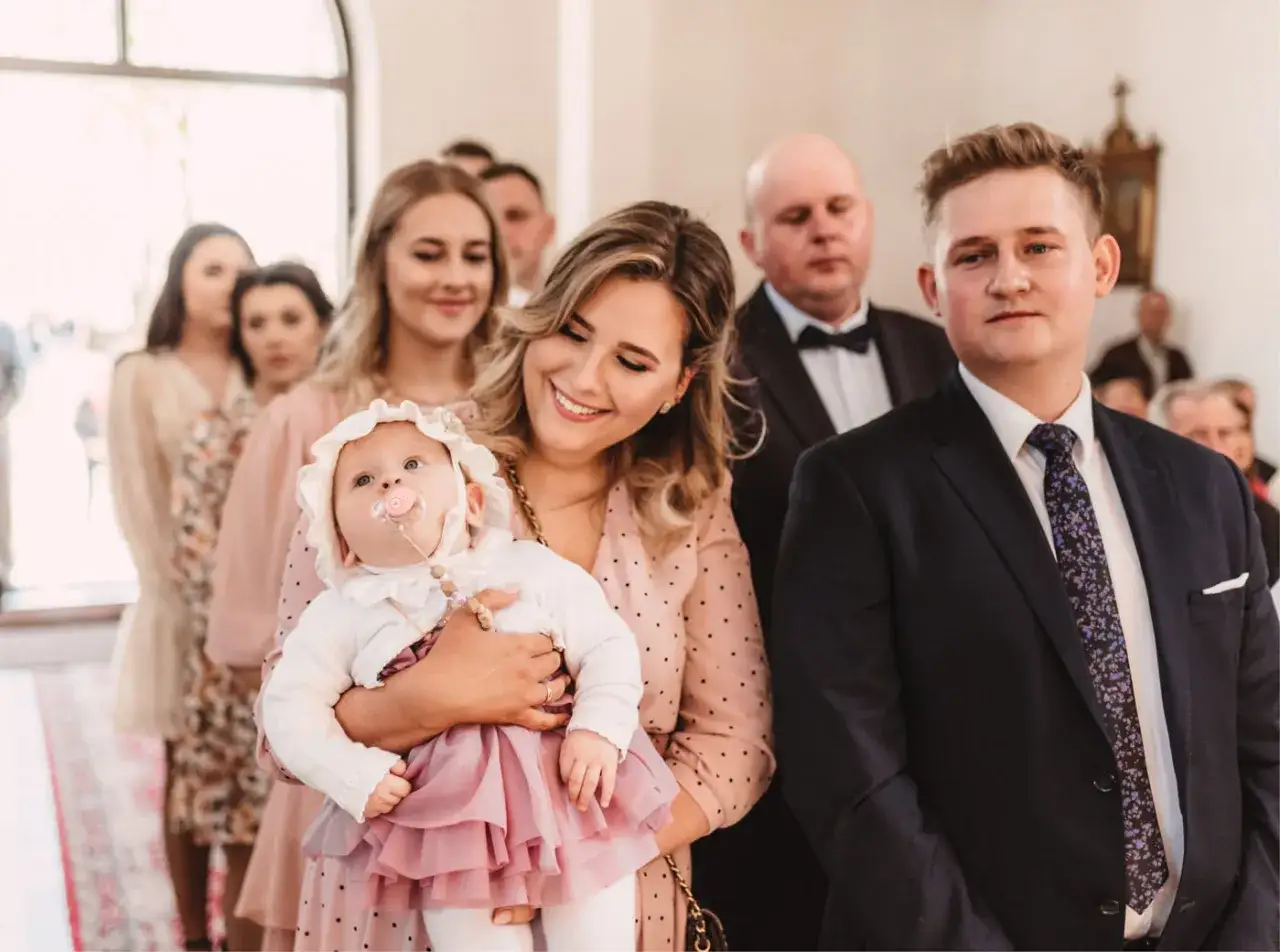 Wzruszające chrzest zdjecia: mama trzyma dziecko w białej czapeczce i różowej sukience, obok dumny ojciec w garniturze.