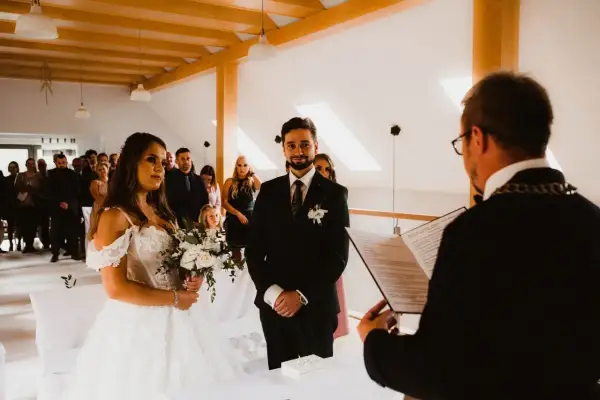 Para młoda podczas ceremonii cywilnej. Panna młoda w białej sukni, pan młody w garniturze. Goście obserwują. Czy po ślubie cywilnym można być chrzestnym?